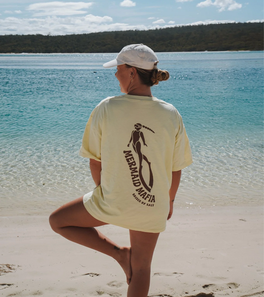 A woman stands on a sandy beach with turquoise water and a forested shoreline in the background, viewed from behind. She is wearing a cream-colored oversized T-shirt with a large black graphic of a woman holding a surfboard and the text 'MERAGLIA MORI' and 'MASSIVE SHAPES'.