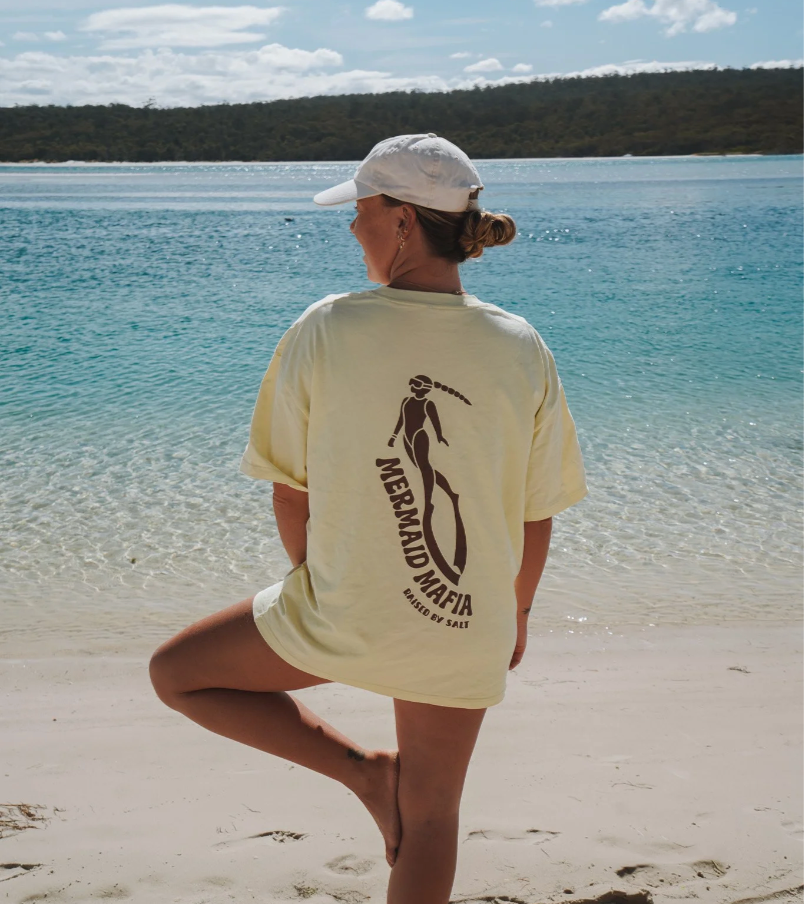 A woman stands on a sandy beach with turquoise water and a forested shoreline in the background, viewed from behind. She is wearing a cream-colored oversized T-shirt with a large black graphic of a woman holding a surfboard and the text 'MERAGLIA MORI' and 'MASSIVE SHAPES'.