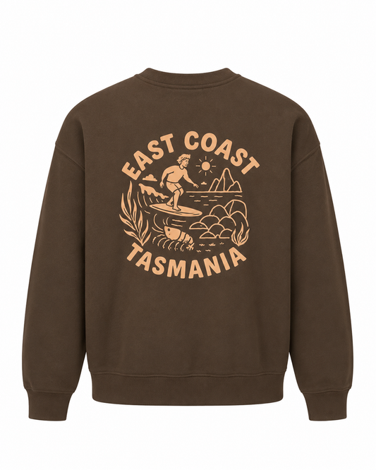 A brown crewneck sweatshirt shown from the back, featuring a large tan graphic print with a surfer, mountains, and the text 'EAST COAST TASMANIA'.