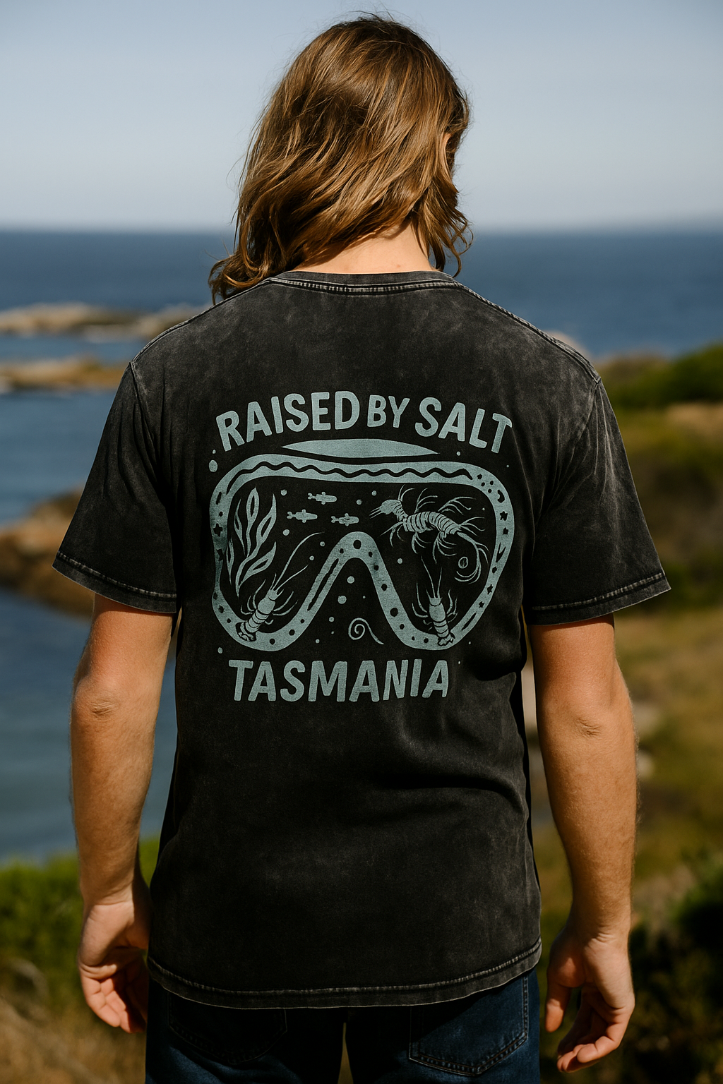 A man viewed from behind wearing a faded black graphic t-shirt with a light blue-green design that reads 'RAISED BY SALT TASMANIA' and features a diving mask illustration. The background shows a scenic coastal landscape with the ocean and cliffs.