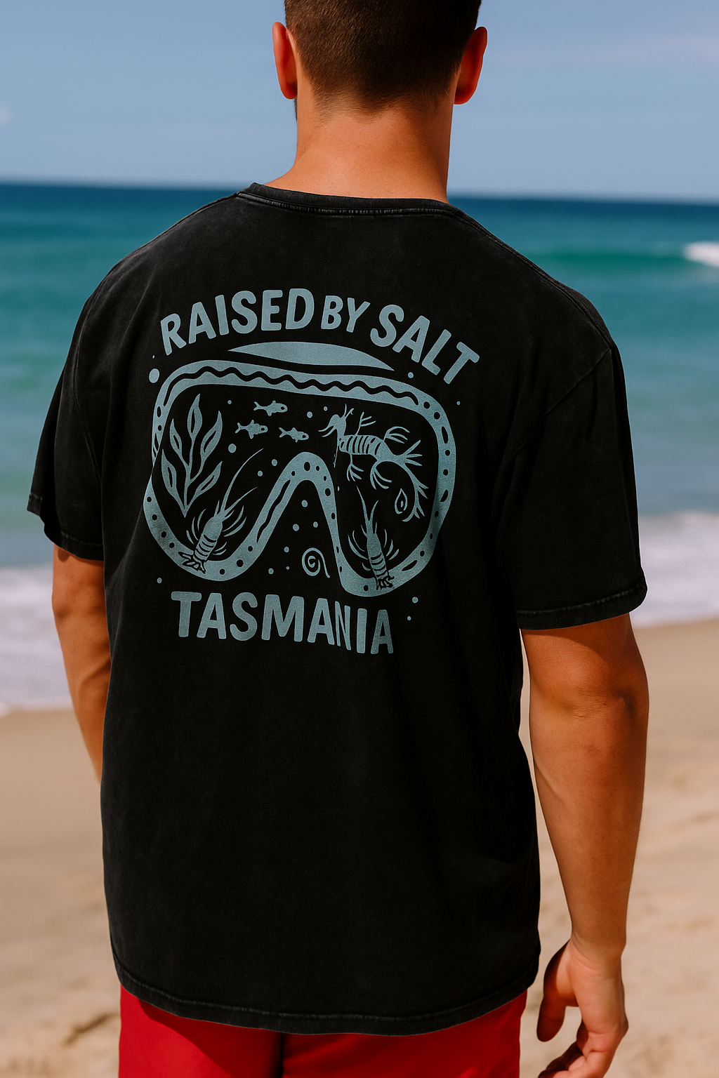 A man standing on a beach with his back to the camera, wearing a black t-shirt with a large light blue graphic on the back that reads 'RAISED BY SALT TASMANIA' around a diving mask design, paired with red shorts.